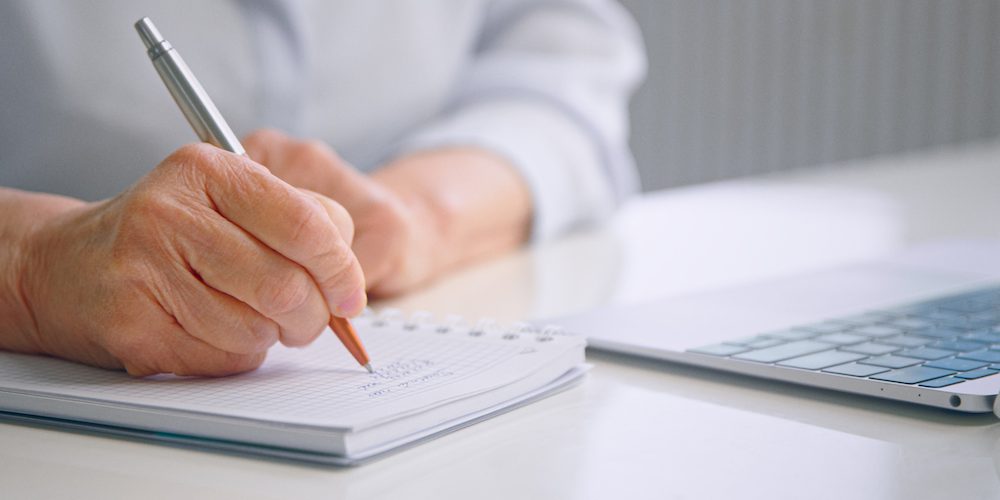 Old lady hand holds grey brown pen and writes notes on paper notebook page near grey laptop on white office table Artificial Intelligence